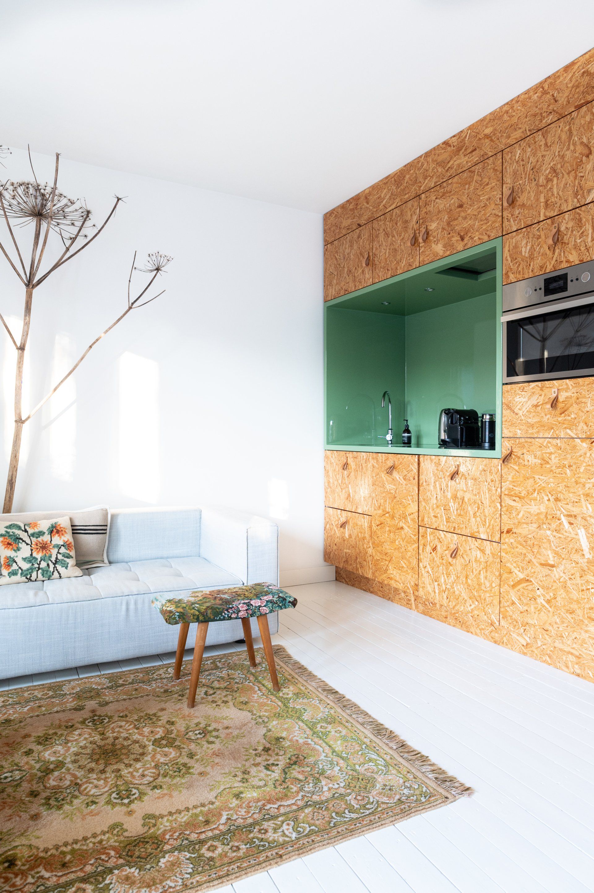 Gezellig, modern appartementinterieur. Een kleine keuken met een groene nis is afgewerkt met houten lambrisering, en er is een kleine bank met een vloerkleed te zien.