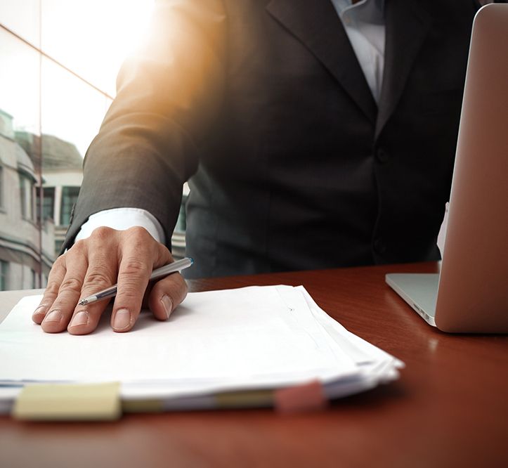 A business professional in a dark suit sits at a desk with a laptop, hand resting on a stack of papers with a pen.