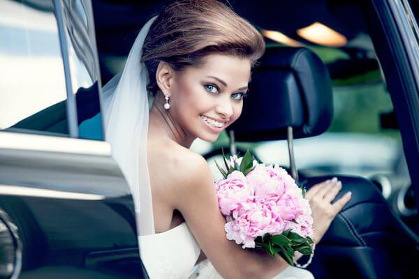 A bride is sitting in the back seat of a car holding a bouquet of pink flowers.