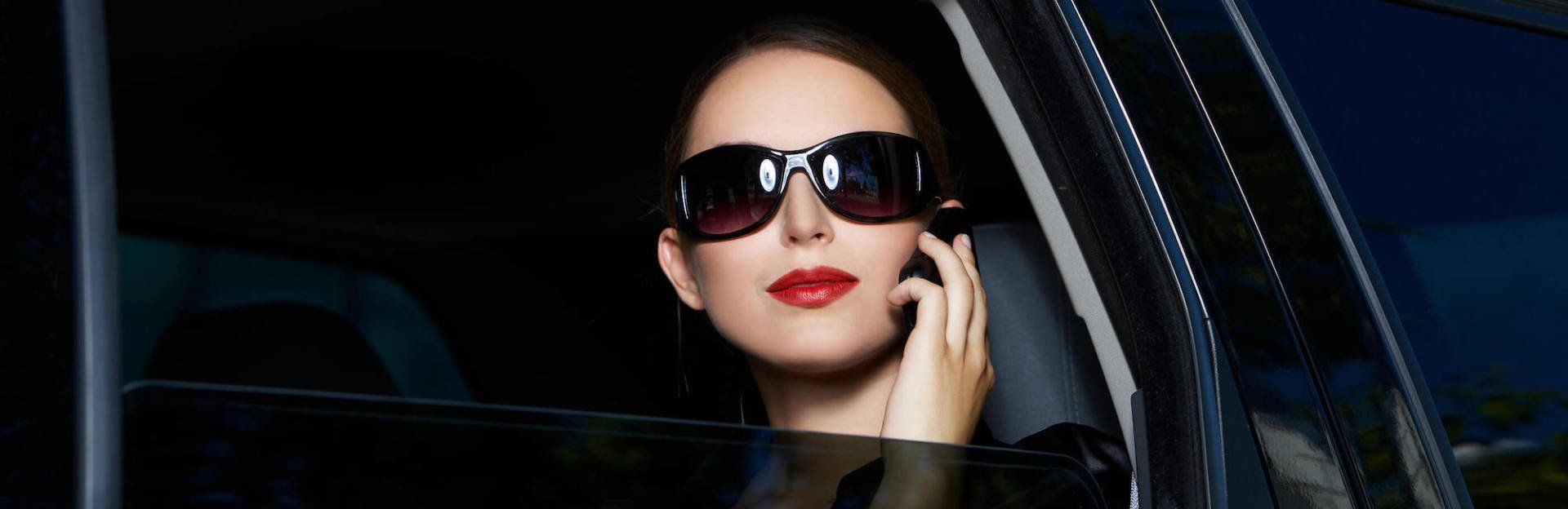 A woman wearing sunglasses is sitting in a car talking on a cell phone.