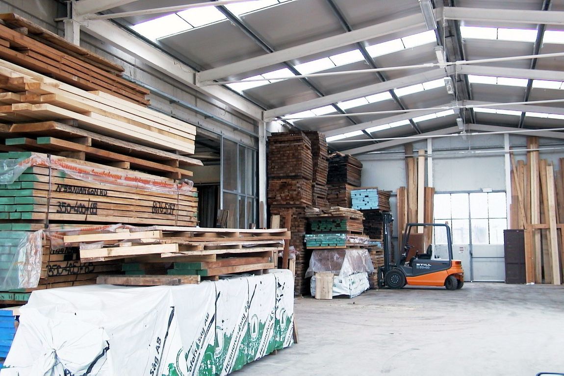 L'interno di un grande magazzino pieno di cataste di legname e assi di legno, con un carrello elevatore arancione sullo sfondo.
