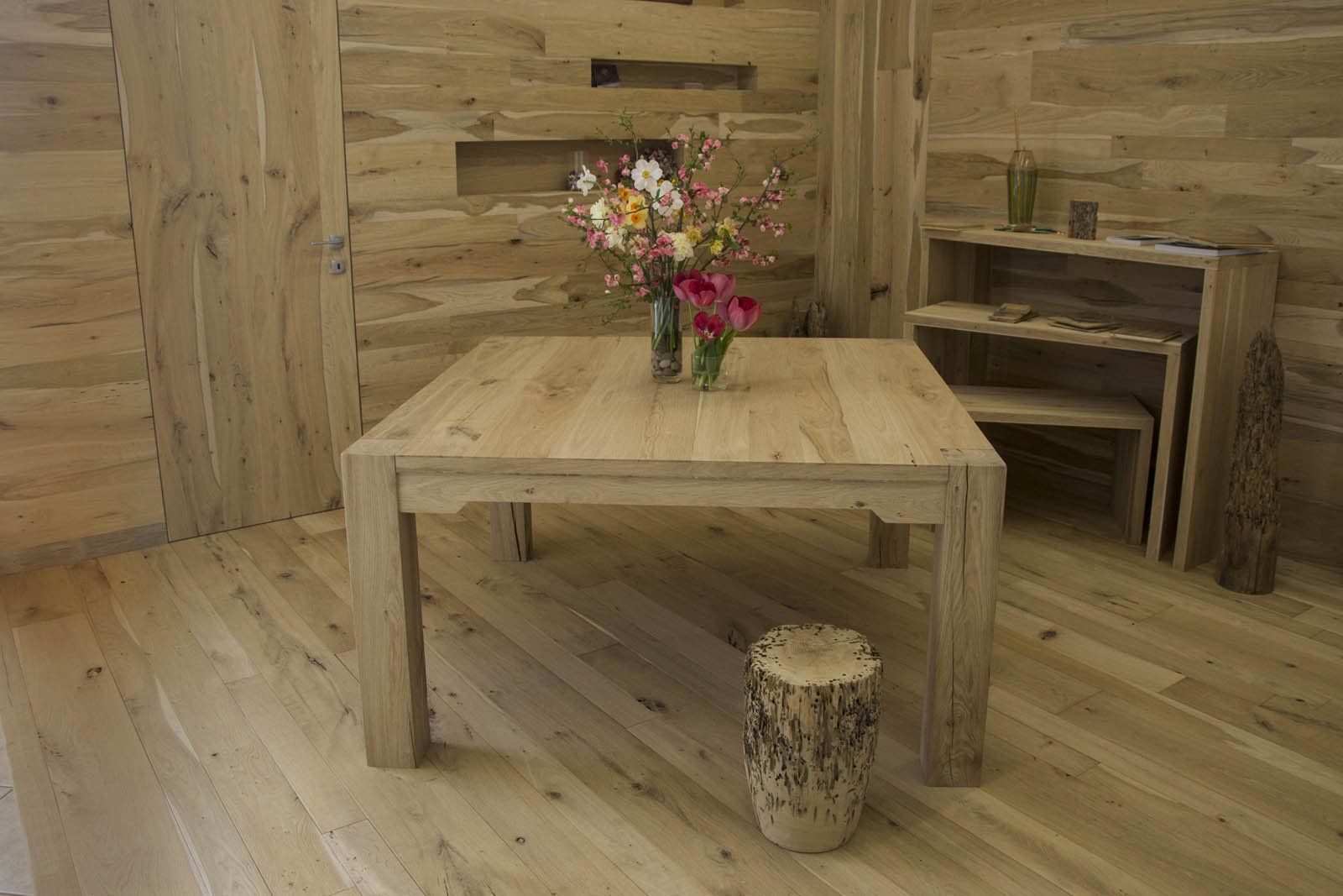 Un tavolo da pranzo quadrato in legno chiaro in una stanza rustica con pareti rivestite in legno coordinato, centrotavola floreale e sgabello in legno massello.