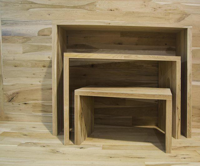 Tre tavolini impilabili in legno chiaro di diverse dimensioni, addossati a una parete rivestita in legno dello stesso colore.