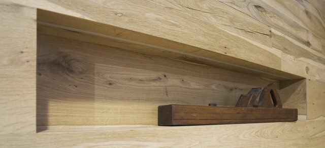 Una pialla a mano in legno scuro è appoggiata su una mensola incassata in una parete rivestita di assi di legno orizzontali di colore chiaro.