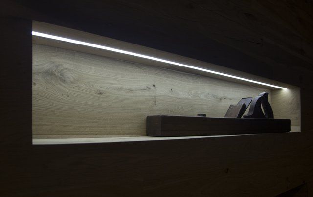 Una mensola a muro in legno illuminata, incassata nel muro, con un oggetto decorativo scuro al centro, in una stanza scarsamente illuminata.
