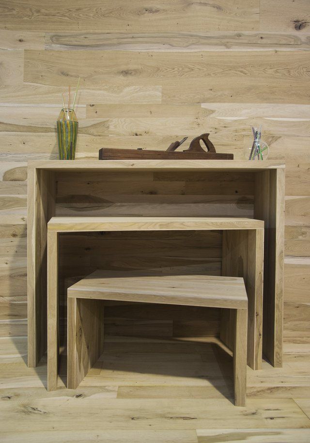 Tre tavolini in legno chiaro, uno dentro l'altro, sono addossati a una parete rivestita di pannelli in legno, con un piccolo piano decorativo sul tavolo più grande.