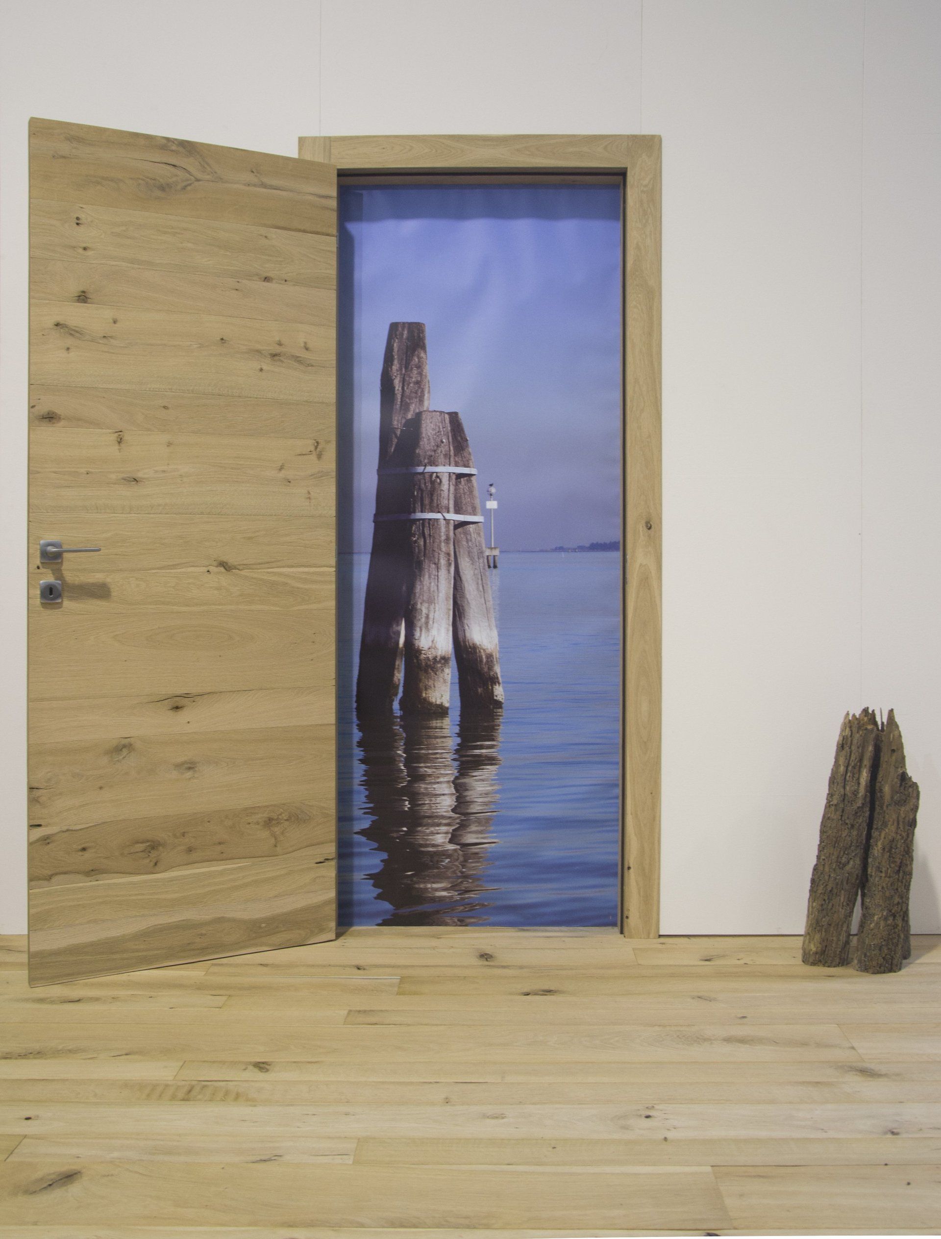 Una porta di legno chiaro è aperta, rivelando un tranquillo panorama acquatico con un palo di legno, incastonato in una parete bianca.