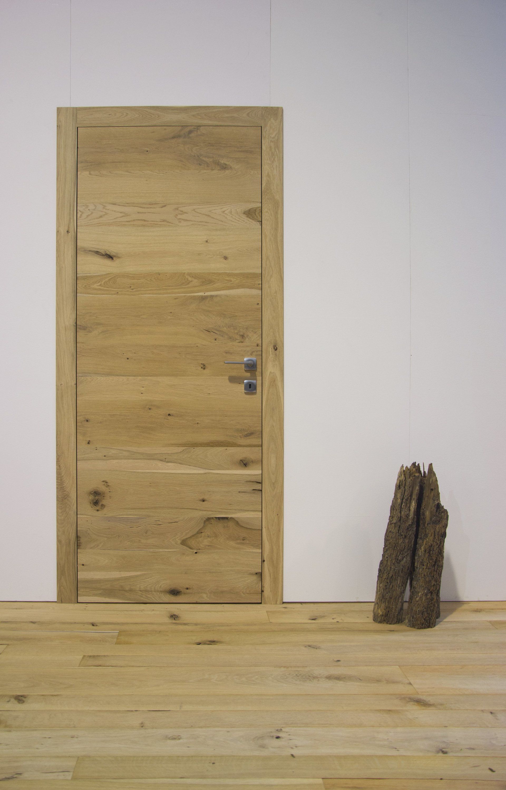 Una porta di legno chiaro incastonata in una parete bianca sopra un pavimento di legno, con un pezzo di corteccia accanto.