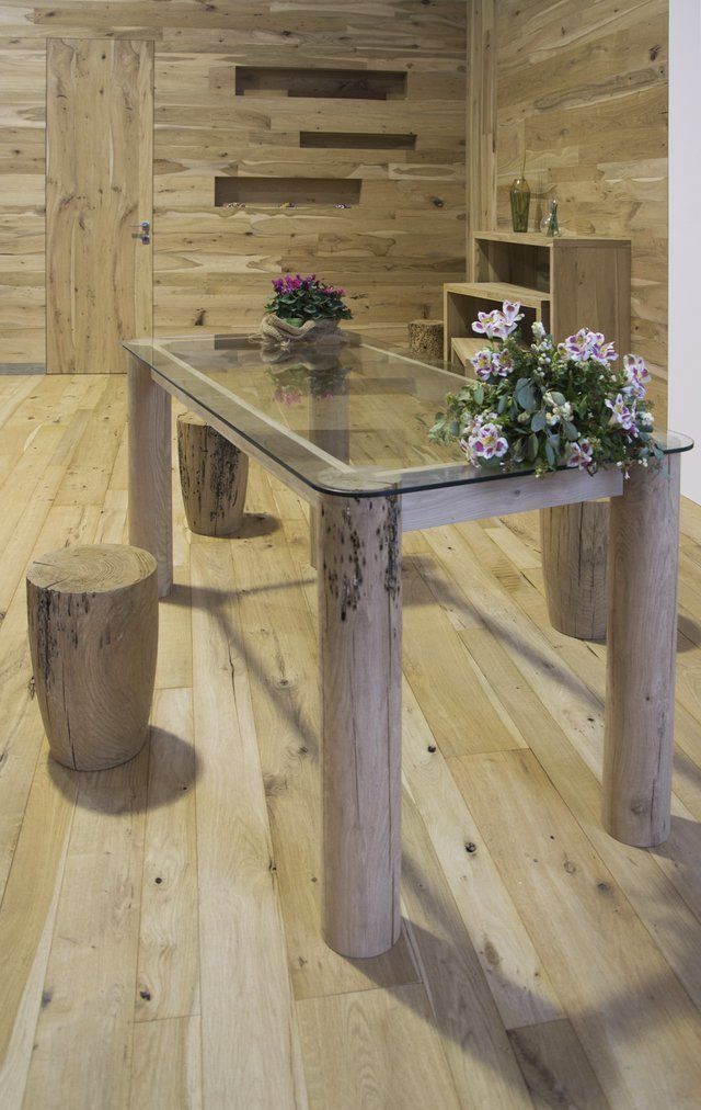 In una stanza rivestita di pannelli in legno, un tavolo da pranzo con piano in vetro, gambe in stile rustico e sgabelli poggia su un pavimento in legno chiaro.