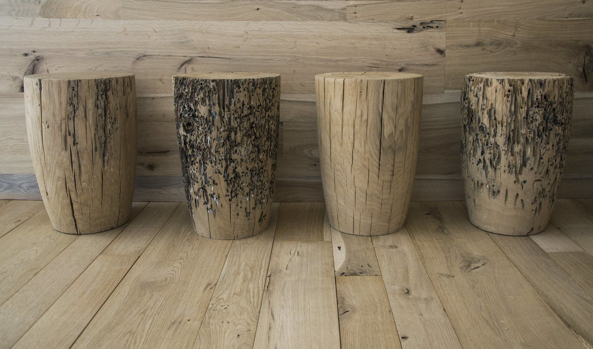 Quattro sgabelli rustici e cilindrici in legno con venature naturali della corteccia, disposti in fila contro una parete rivestita di pannelli di legno chiaro.