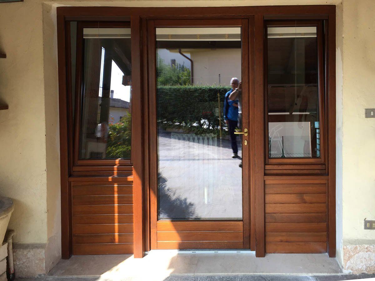 Una porta d'ingresso in legno con pannelli in vetro, affiancata da due finestre laterali identiche con pannelli orizzontali in legno nella parte inferiore.
