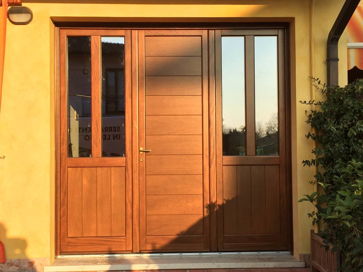 Una porta d'ingresso in legno marrone con pannelli laterali dotati di finestre verticali, su uno sfondo giallo.