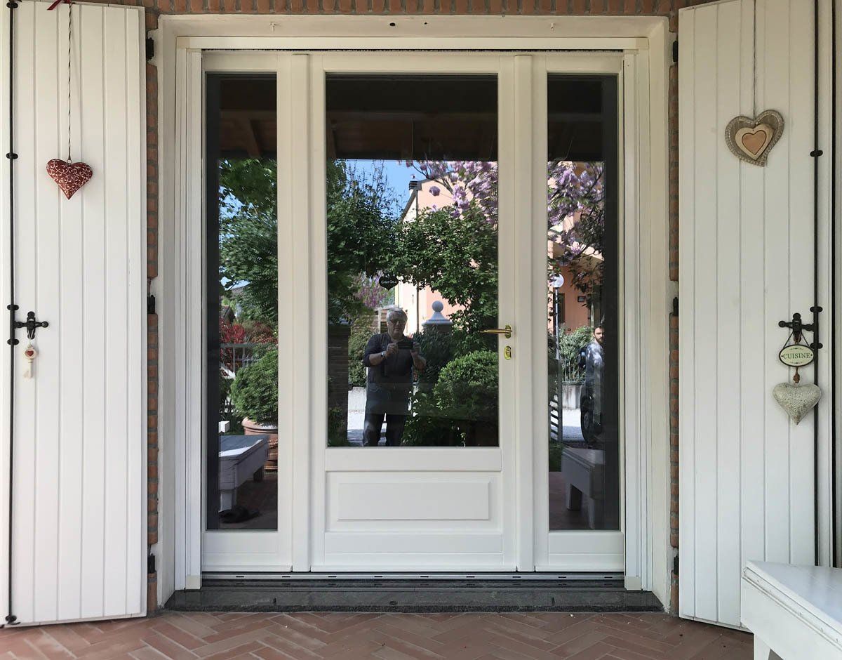 Una porta d'ingresso bianca con pannelli laterali, fiancheggiata da persiane in legno, con una persona riflessa nel vetro.