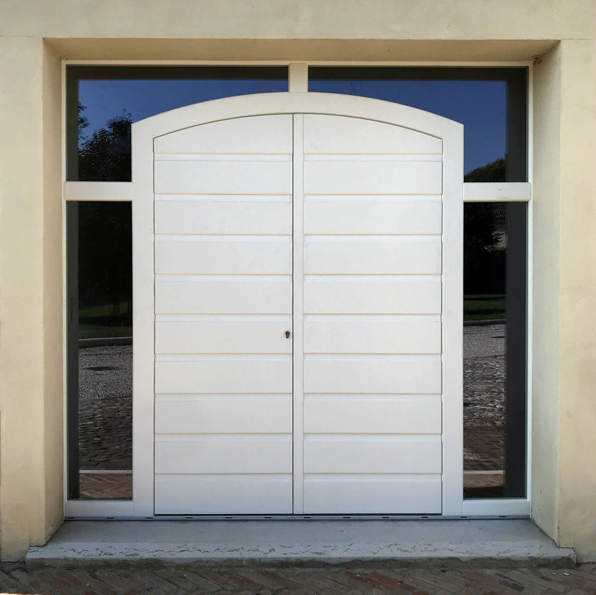 Doppie porte bianche con pannellatura orizzontale e parte superiore arrotondata, inserite in una cornice di pannelli di vetro scuro su una parete beige.