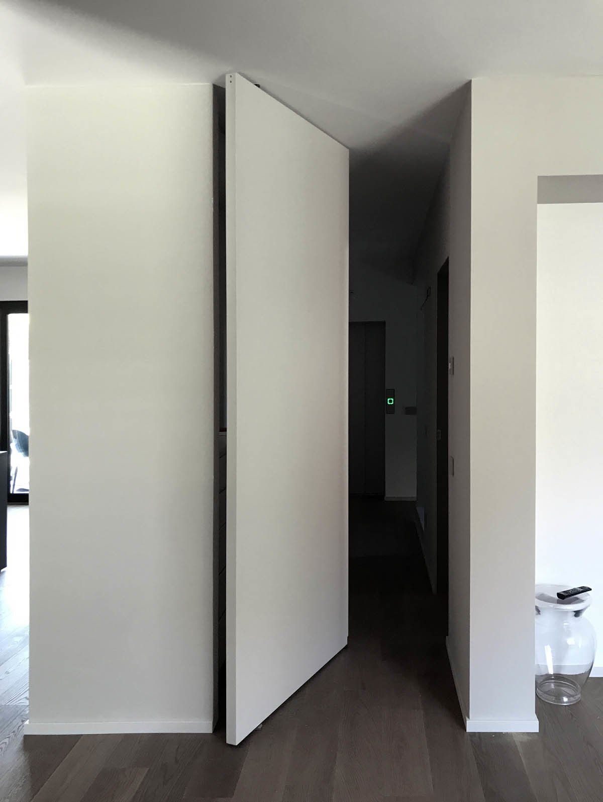 In un corridoio moderno e minimalista con pavimento in legno, una porta a bilico bianca, senza telaio e a tutta altezza, rimane leggermente socchiusa.