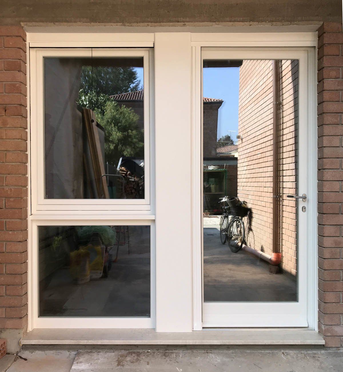 Una finestra con cornice bianca e due vetri sovrapposti, accanto a una porta bianca abbinata, inserita in un muro esterno di mattoni.