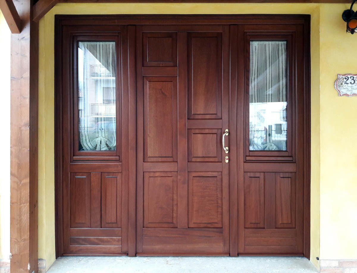 Una porta d'ingresso in legno scuro con due finestre laterali su una parete gialla, caratterizzata da una maniglia in ottone e dettagli a pannello rettangolare.