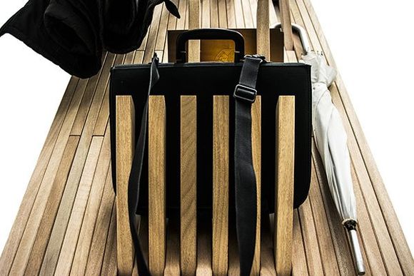 Una valigetta nera è appoggiata in verticale tra le doghe di legno di una panchina, con una borsa nera e un ombrello bianco nelle vicinanze.