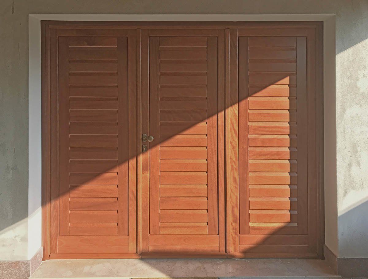 Un set di tre porte a persiana in legno installate in una parete chiara, parzialmente in ombra.