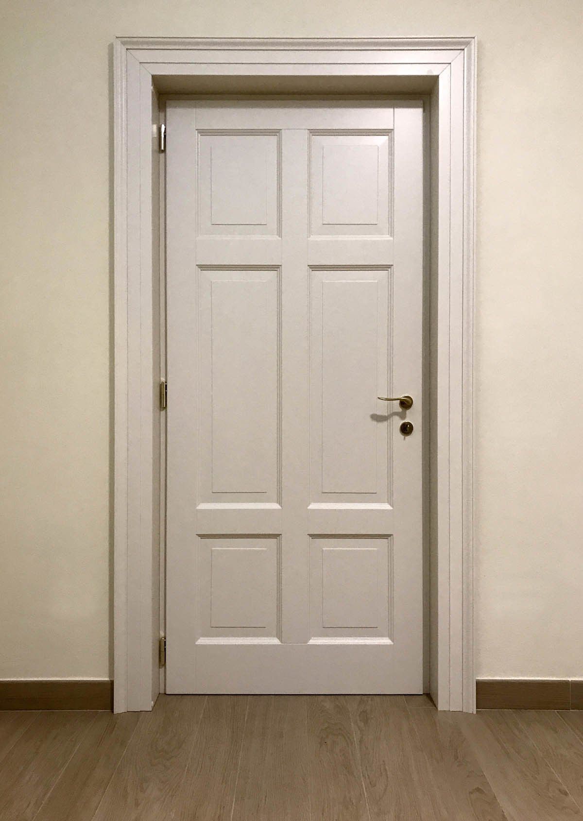 Una porta interna bianca a sei pannelli, con maniglia dorata e modanatura bianca, su uno sfondo di parete chiara e pavimento in legno.