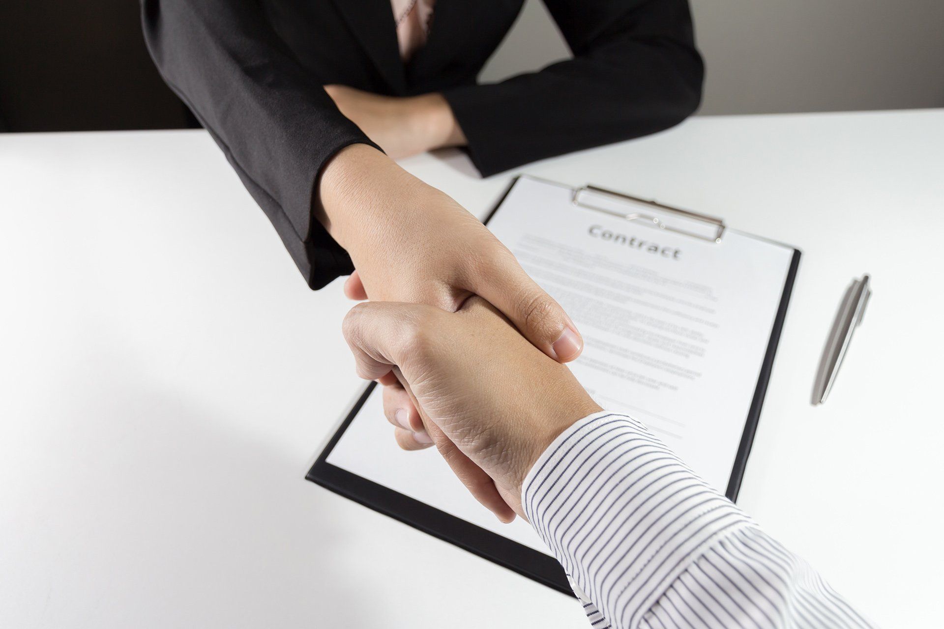 Two people shaking hands over a contract on a white table.