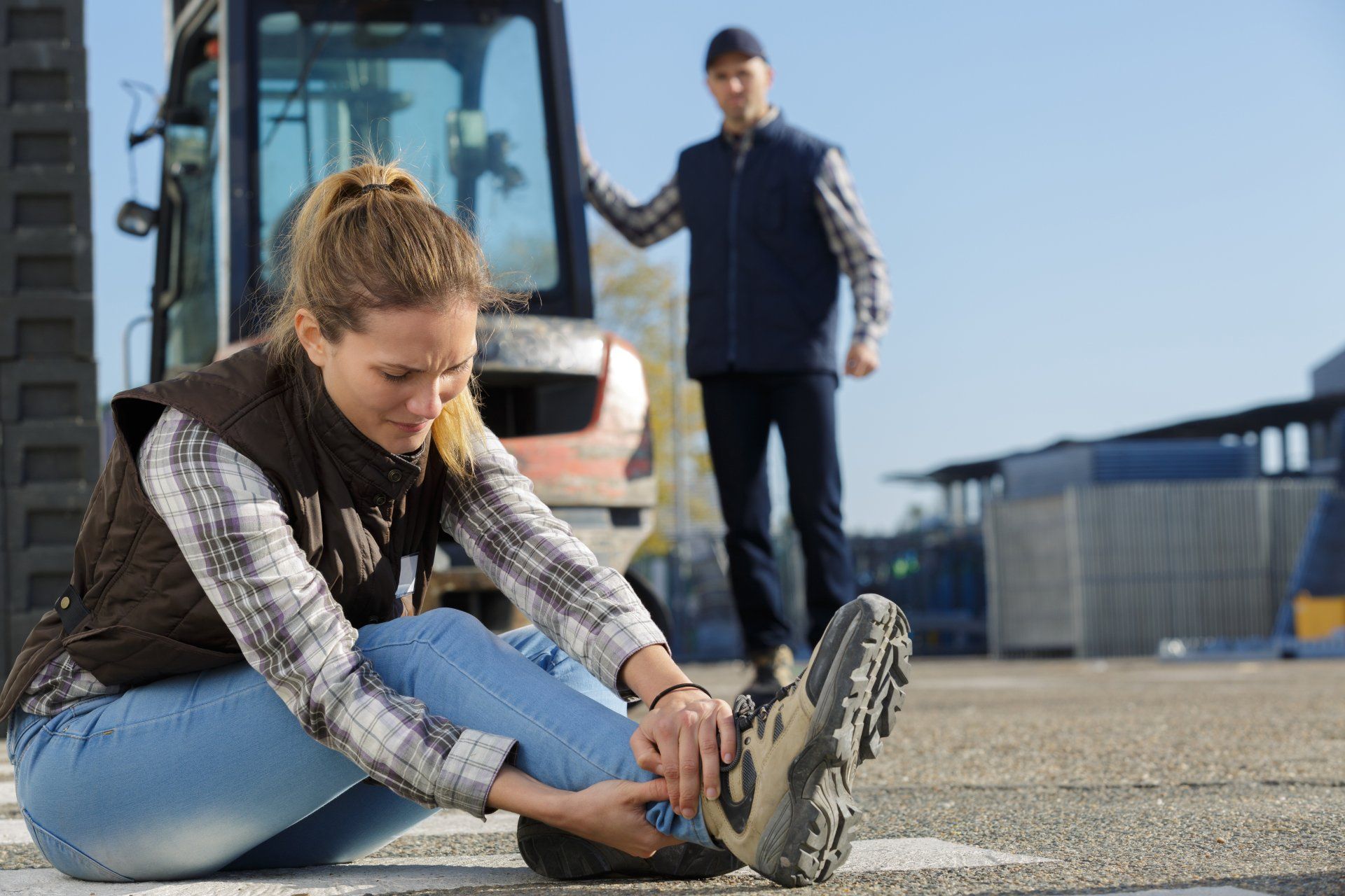 Woman clutching her ankle after a possible accident near a construction vehicle, man in background.