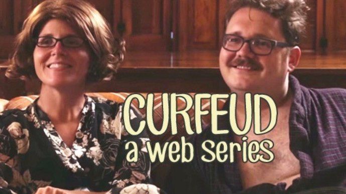 Two people in wigs on a couch; the text