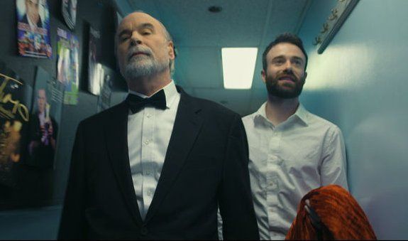 Man in tuxedo and another man with beard walking in a hallway; red wig visible.