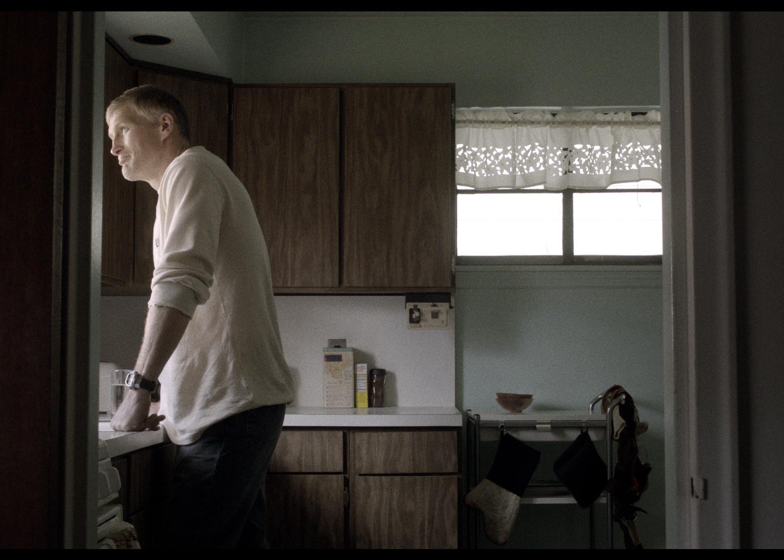 Man in a white shirt in a kitchen with brown cabinets, looking toward a window.