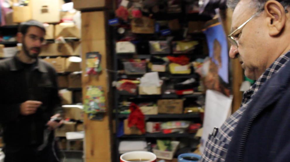Man in foreground, focus on side profile, shop cluttered with shelves and another person behind.