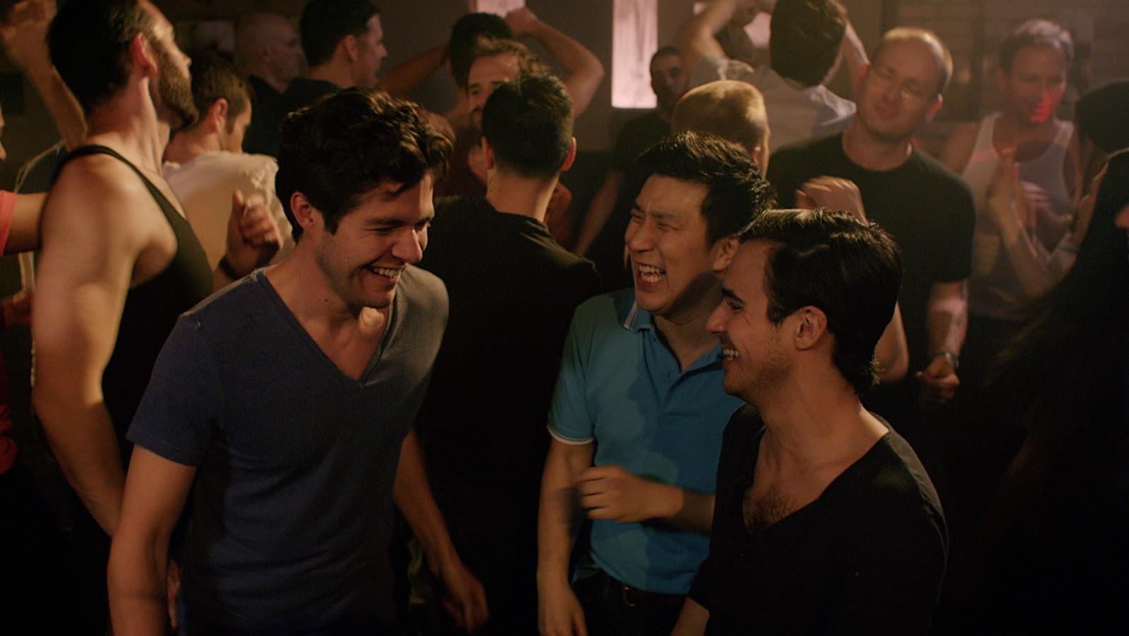 Men dancing in a crowded nightclub, smiling and laughing. Dark setting, people wearing casual clothes.