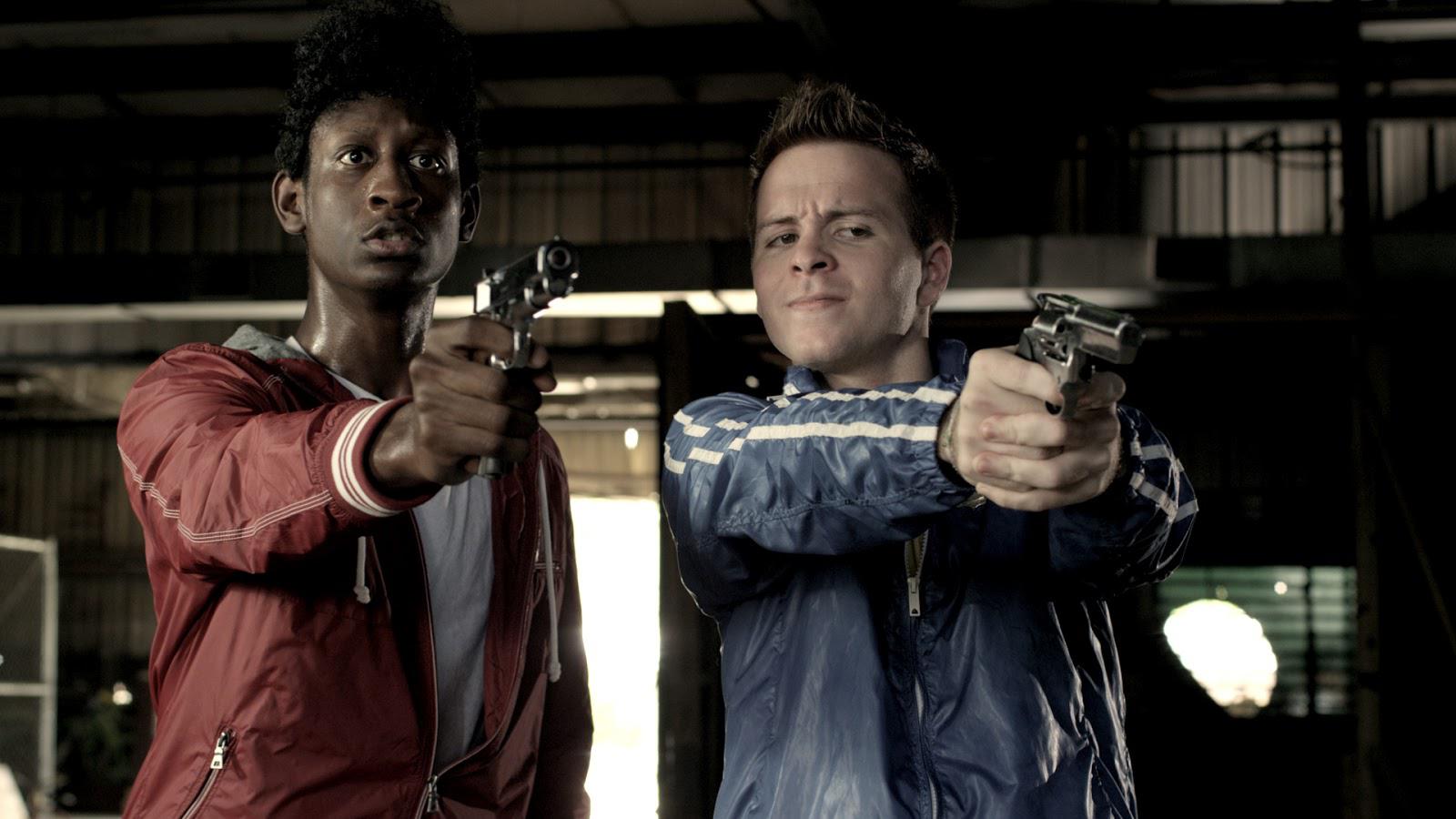 Two young men, one black, one white, aiming pistols in a dark industrial setting.