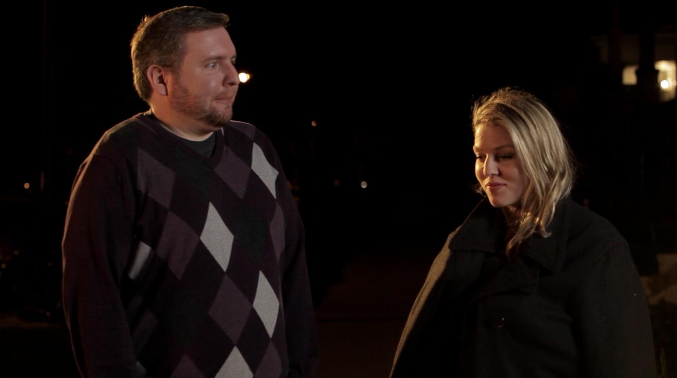 Man and woman outside at night. Man in argyle sweater looks to the side, woman smiles down.
