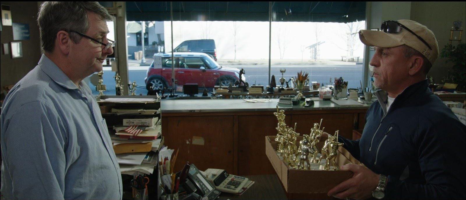 Two men in shop, one holding box of trophies. Red car visible through window.
