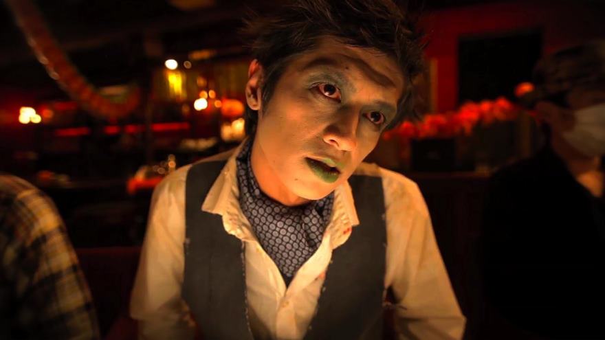 A person with ghoulish makeup stares forward in a dim, restaurant-like setting.