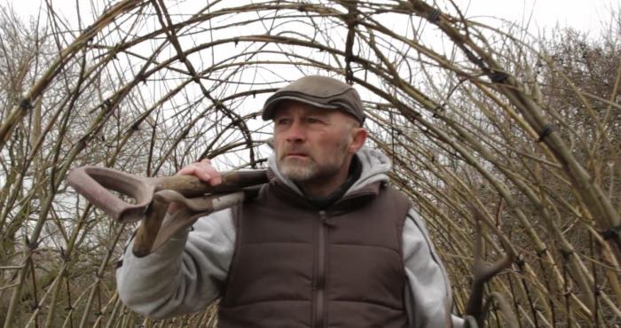 Man with shovel inside willow archway, wearing cap and vest, looking to the side.