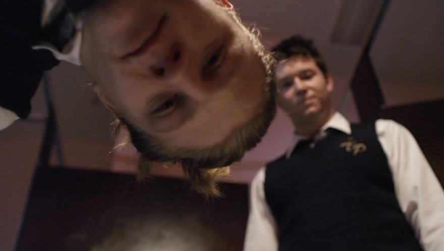 Two men looking down at something; one is upside down. Indoor setting, one wears a vest.