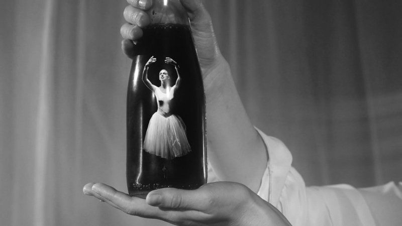 Black and white photo of hand holding a dark glass bottle, a ballerina is reflected inside the bottle.
