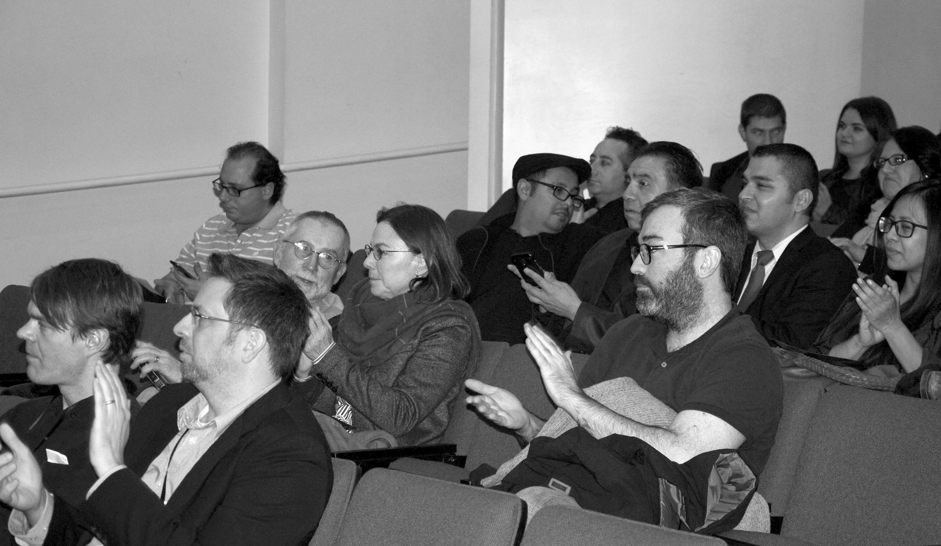 Audience applauds in a brightly lit auditorium. Men and women, diverse in age and style, clap with smiles.