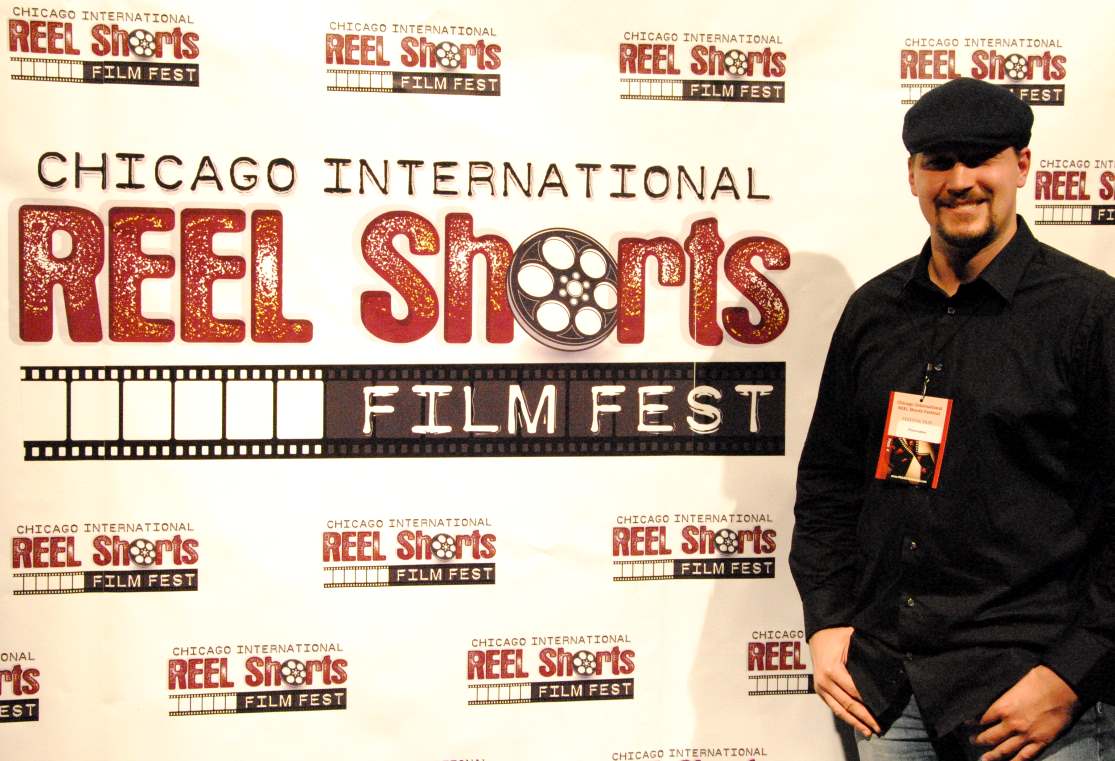 Man stands next to a Chicago Reel Shorts Film Fest backdrop. He wears a dark shirt and cap.