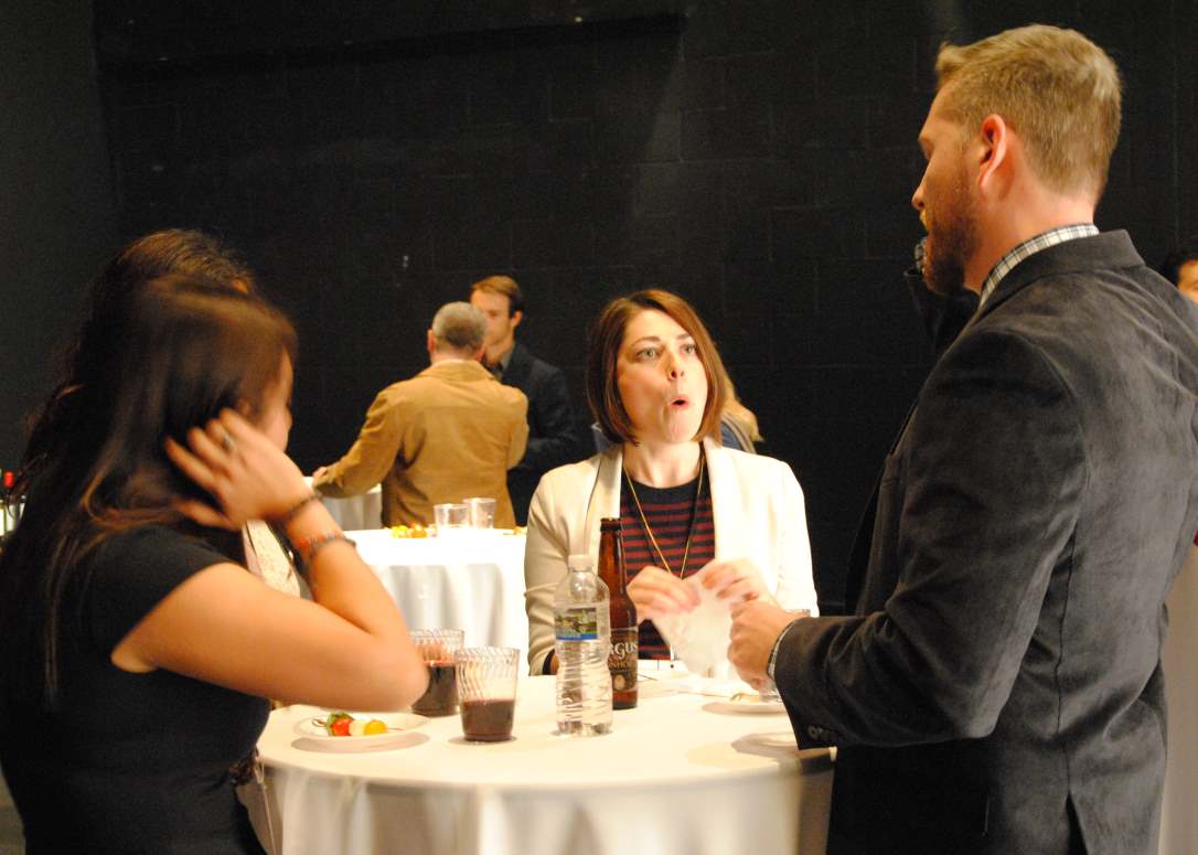 People at a gathering around a small table. Woman in white jacket talks animatedly. Man in blazer listens.