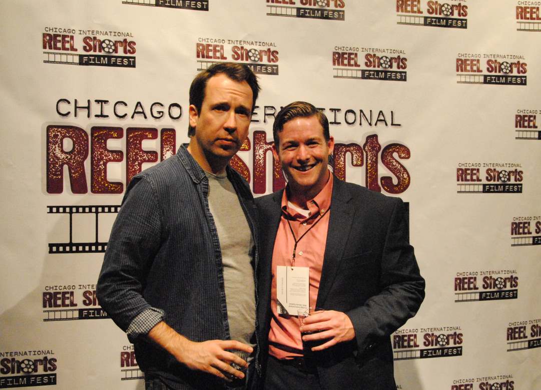 Two men pose at the Chicago Reel Shorts film festival. One wears a denim jacket, the other a blazer.