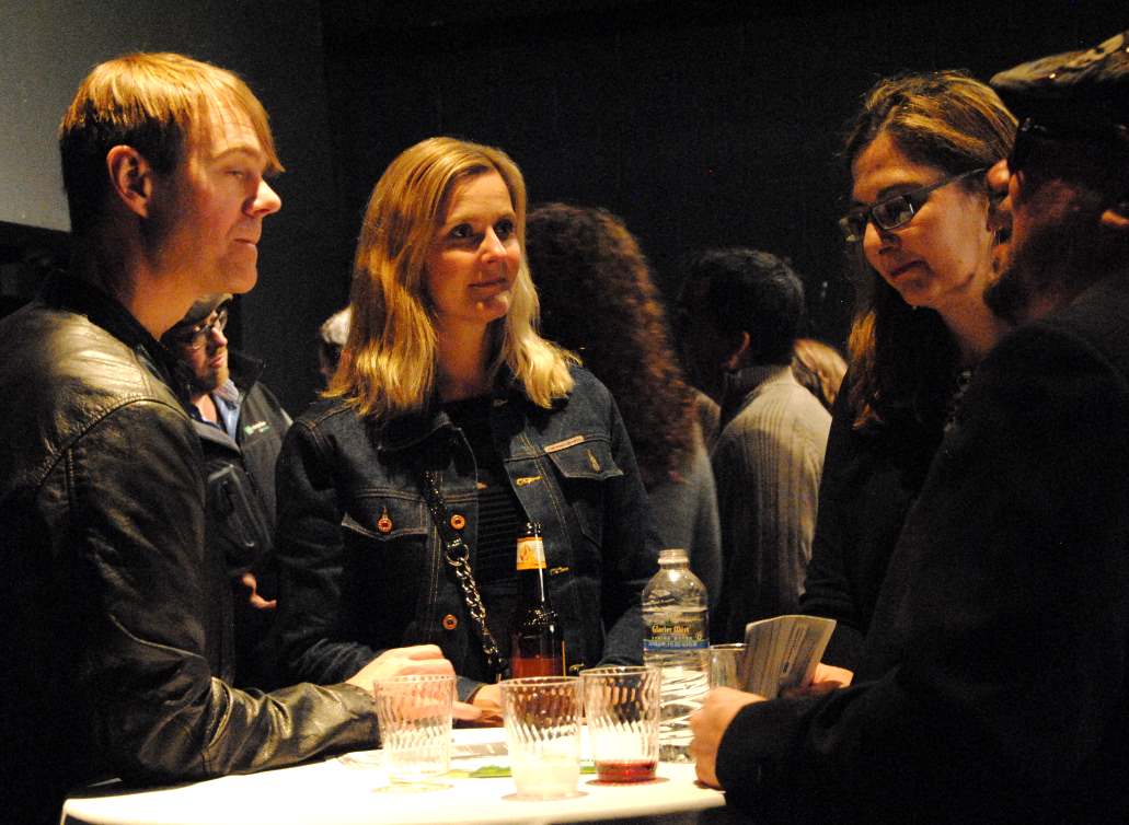 People talking at a table in a dimly lit room. Woman in denim jacket, man in leather jacket.