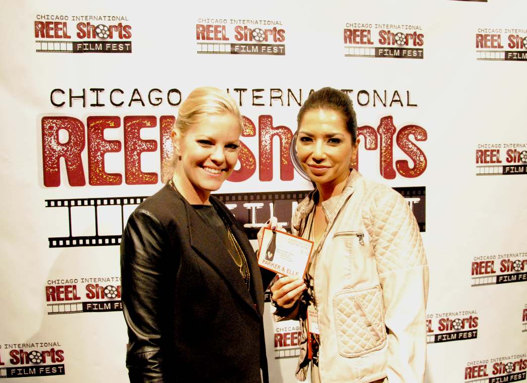 Two women smiling at a film festival; one holds a card. Chicago Reel Shorts backdrop.