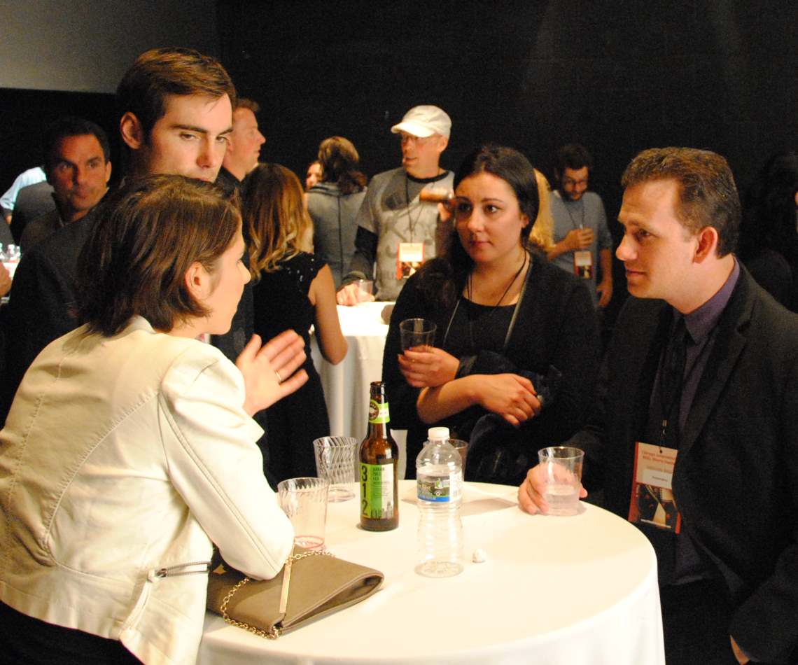 People at a networking event, conversing around a table with drinks. Low lighting.
