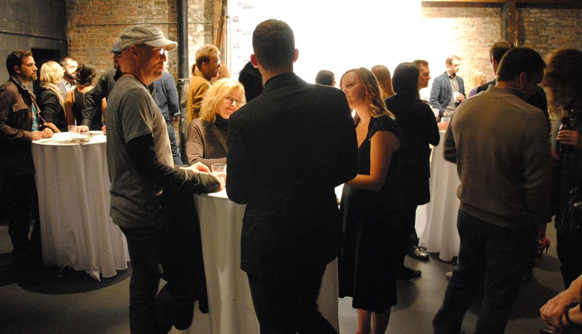 People at a social event, standing around white tables, talking. Mostly dressed in dark clothing, indoors.