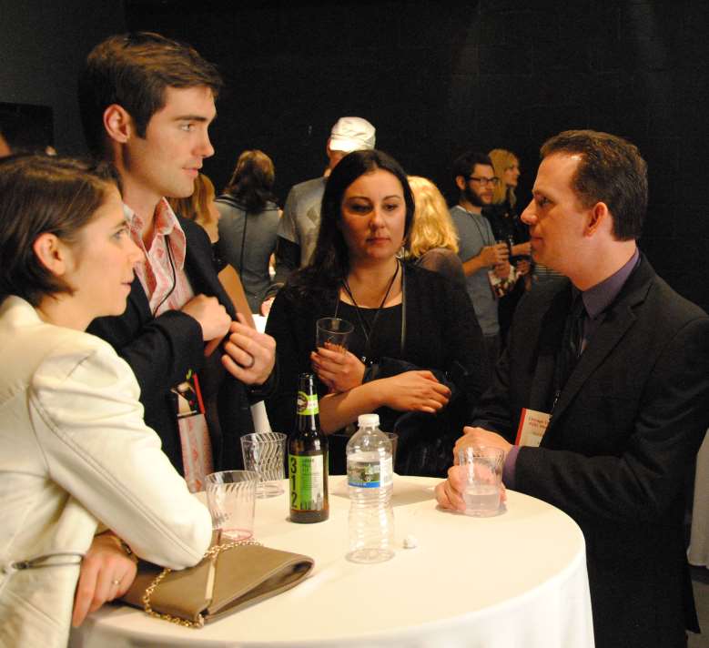 People conversing around a small table, holding drinks. Attendees at an event with neutral backdrop.