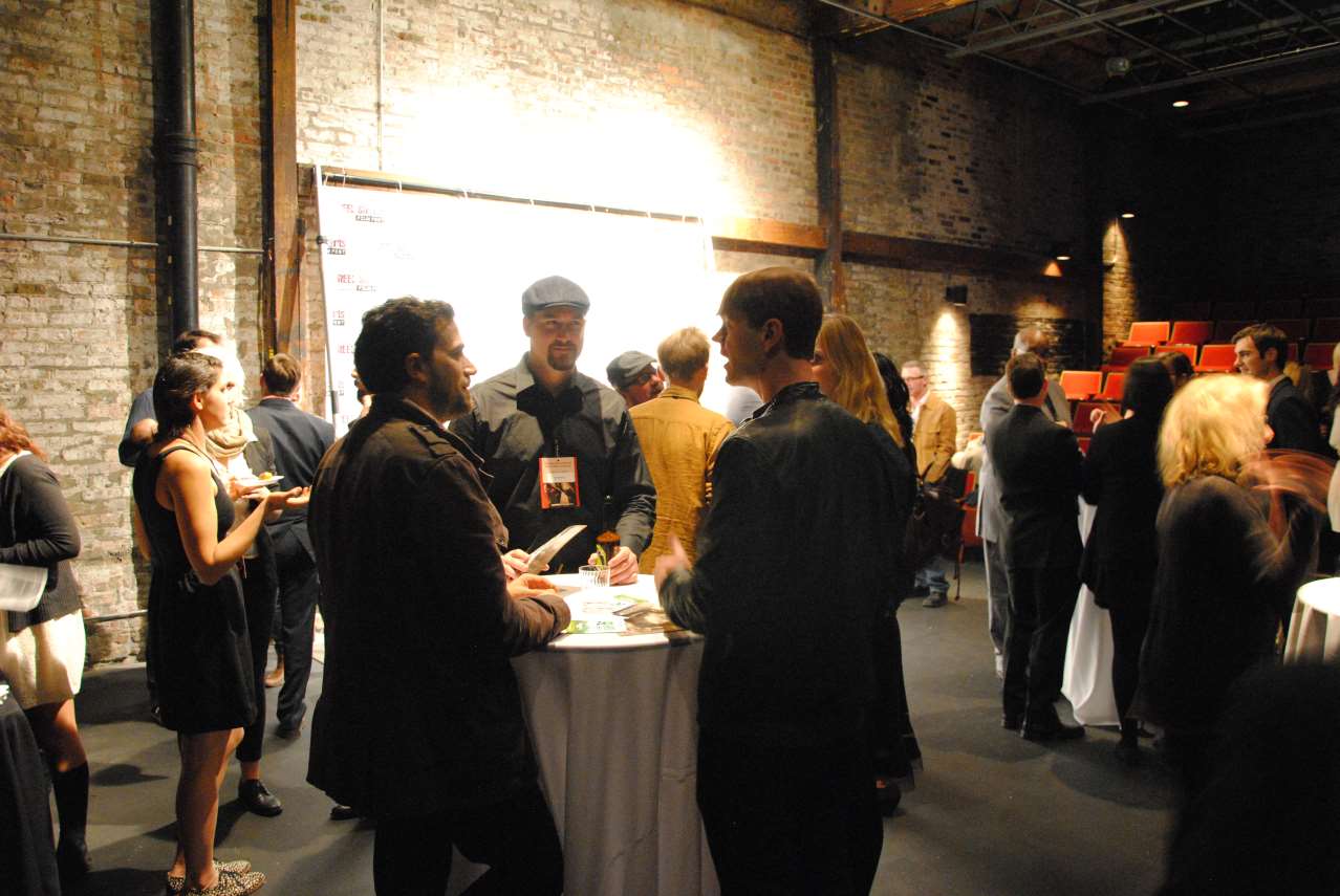 People mingle at an event in a dimly lit industrial space. They stand near a table and walls.