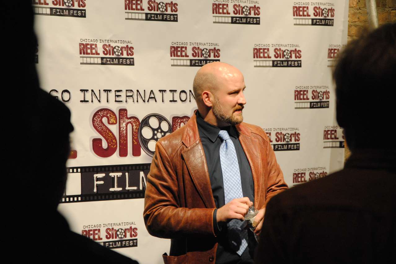 Man with shaved head wearing leather jacket, blue tie, standing at a film festival, speaking.
