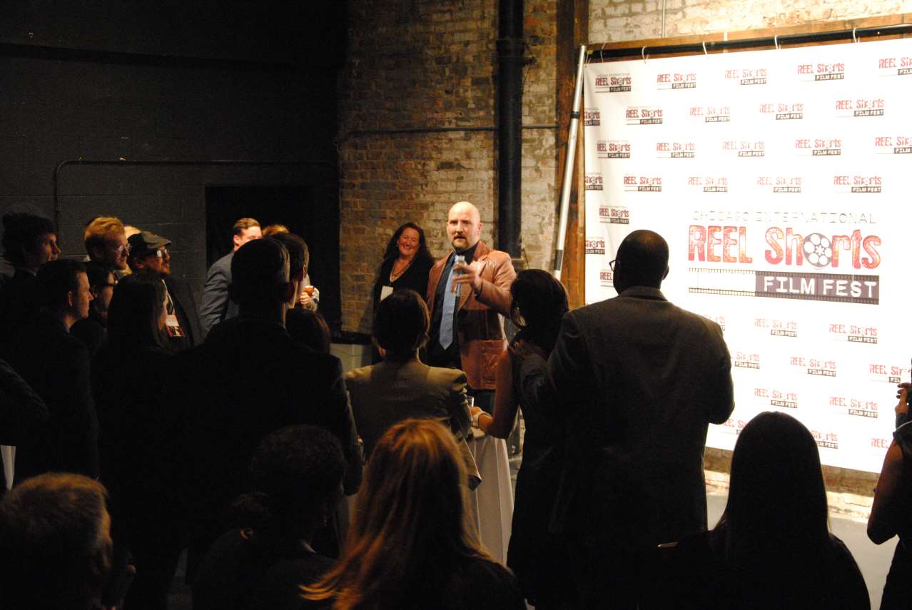 Man speaking at a film festival, surrounded by an audience in a dimly lit industrial space.